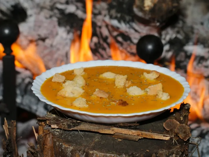 Une assiette creuse avec une soupe orangée et des croutons de pain est présentée devant un feu de cheminée allumé
